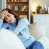 Woman sitting on couch with hand on forehead showing fatigue and headache symptoms of untreated iron deficiency