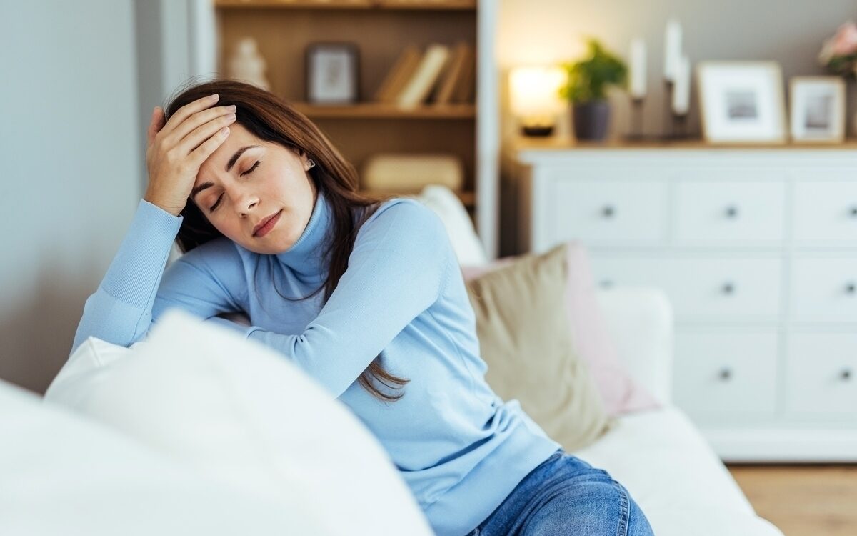 Woman sitting on couch with hand on forehead showing fatigue and headache symptoms of untreated iron deficiency