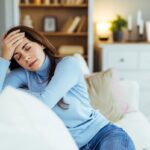 Woman sitting on couch with hand on forehead showing fatigue and headache symptoms of untreated iron deficiency
